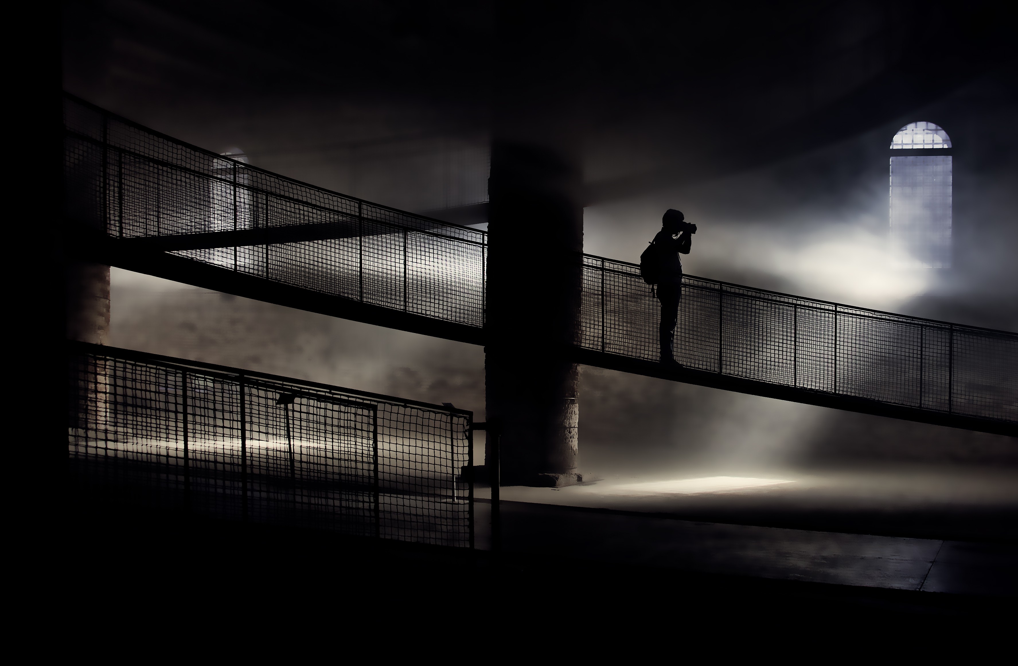 Silhouetted photographer on an elevated walkway in a dark, misty hall.
