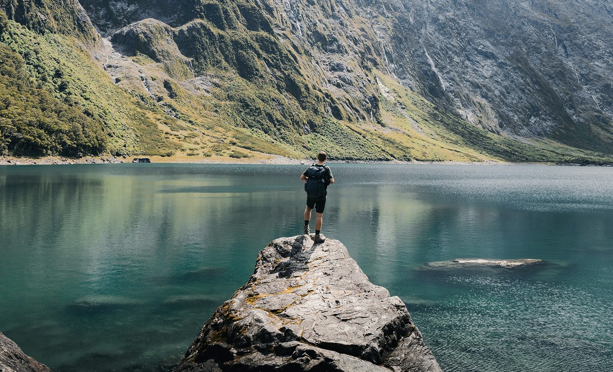 A man stands on a rock, looking at a calm lake surrounded by mountains.