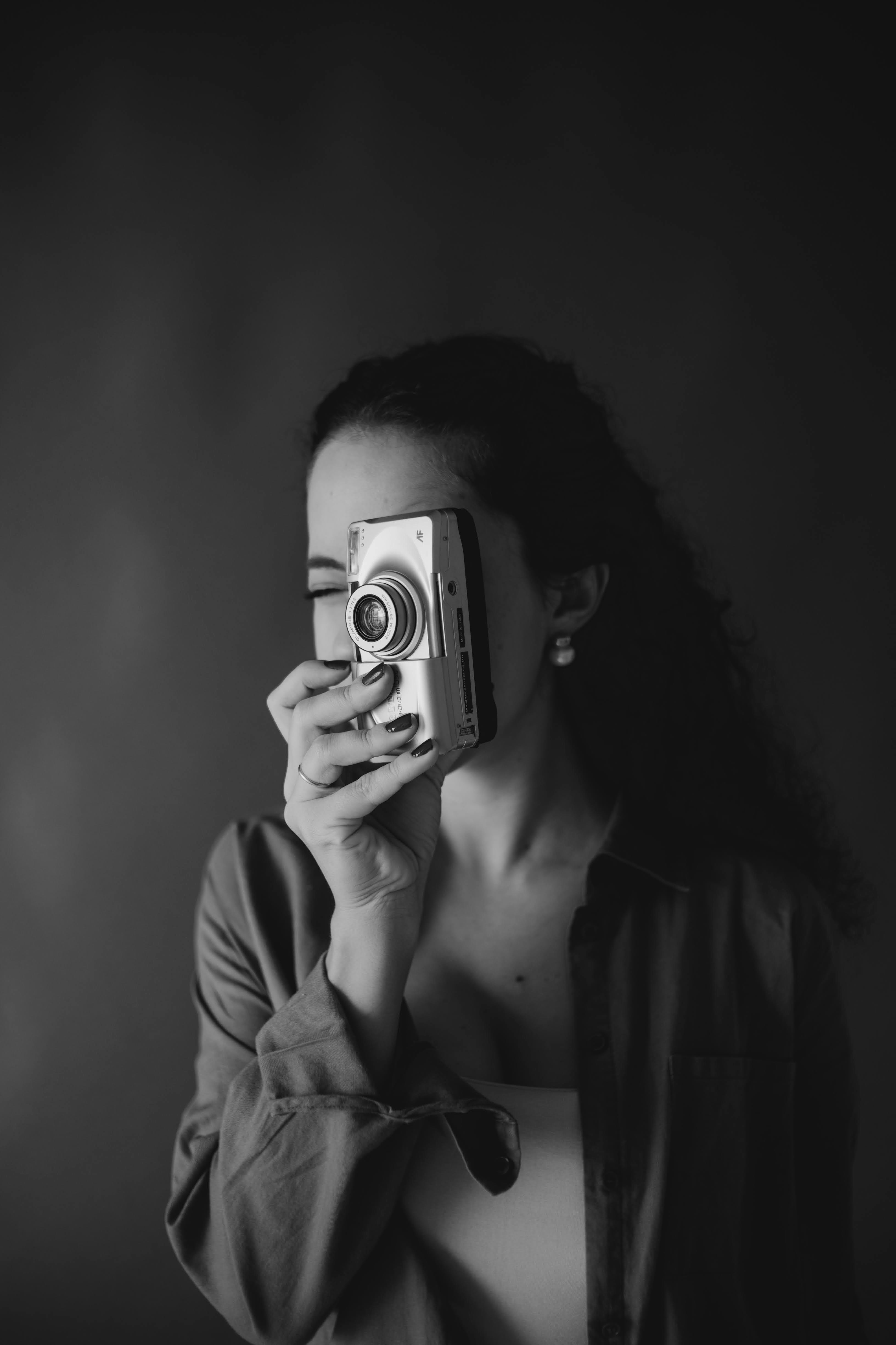 Black and white portrait of a woman with curly hair holding a compact camera in front of her face, wearing a loose shirt and pearl earrings.