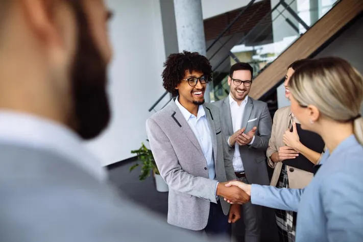 Group of professionals in business attire standing in a room, smiling and shaking hands, reflecting a positive and successful meeting. The setting is formal, with a collaborative atmosphere and a focus on networking or agreement.