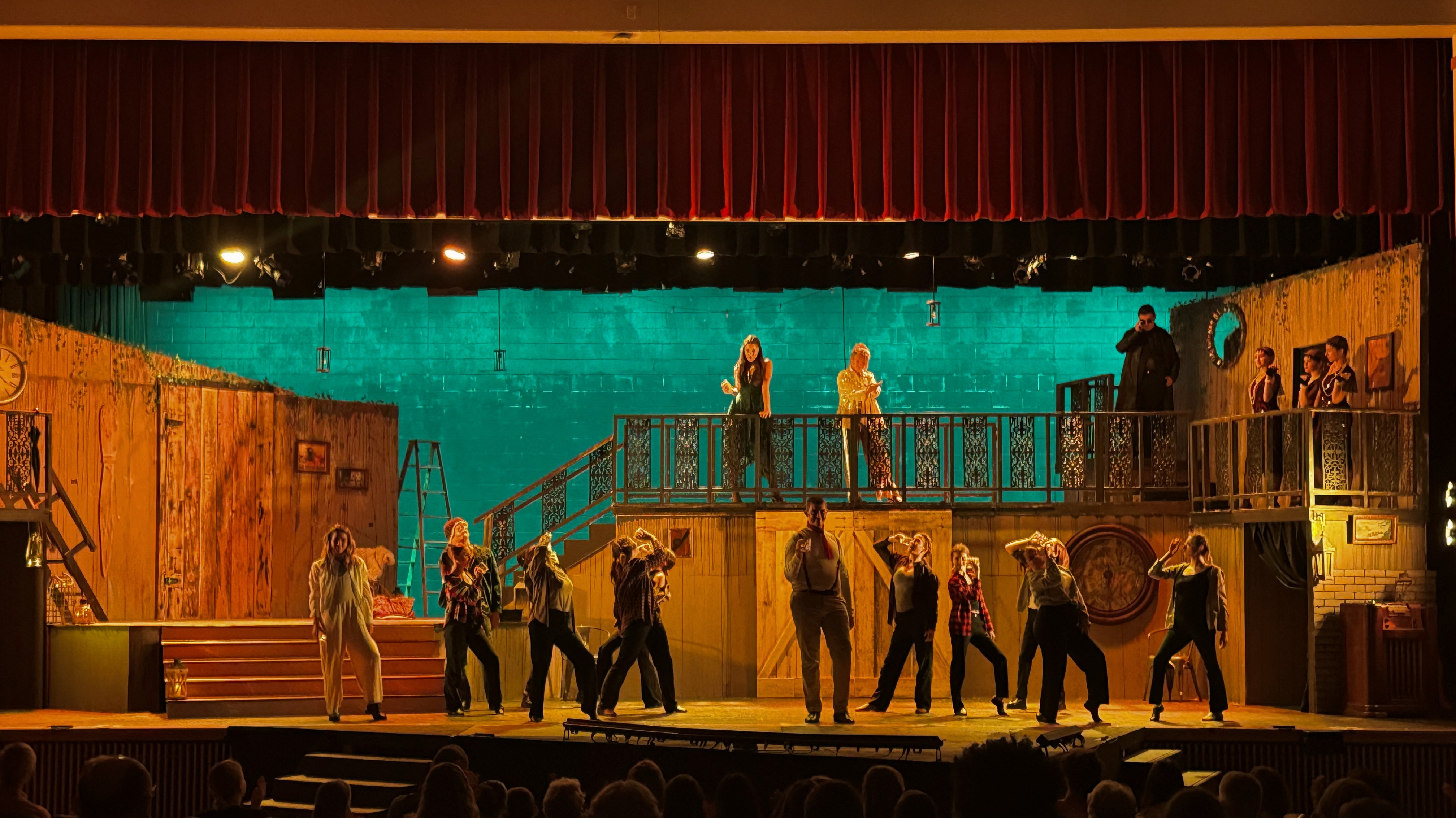 A vibrant theatrical performance on stage features actors in dynamic poses on several layers of a set. Behind them is an almost-green CYC. They are illuminated with warm tones from the side and top.