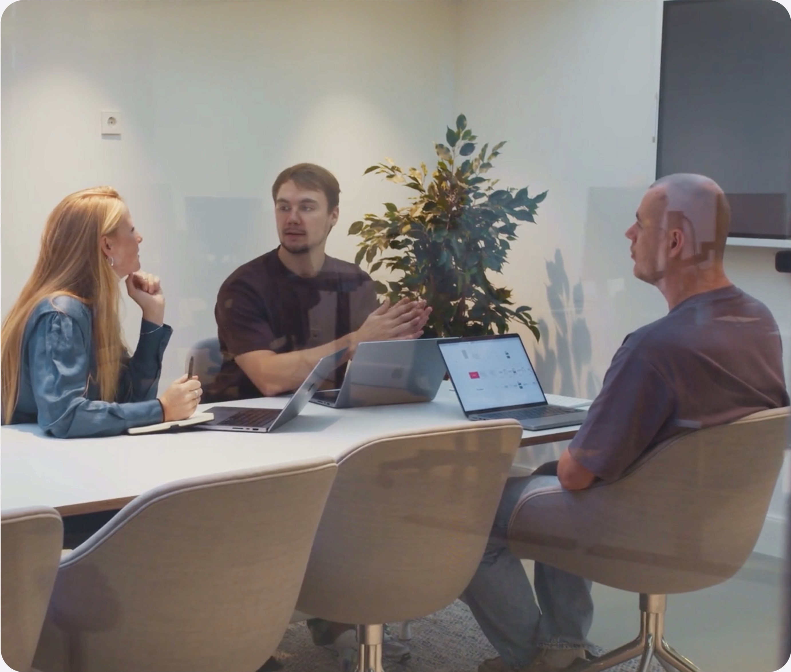 Three people engage in conversation at a conference table with laptops and a plant in the background.
