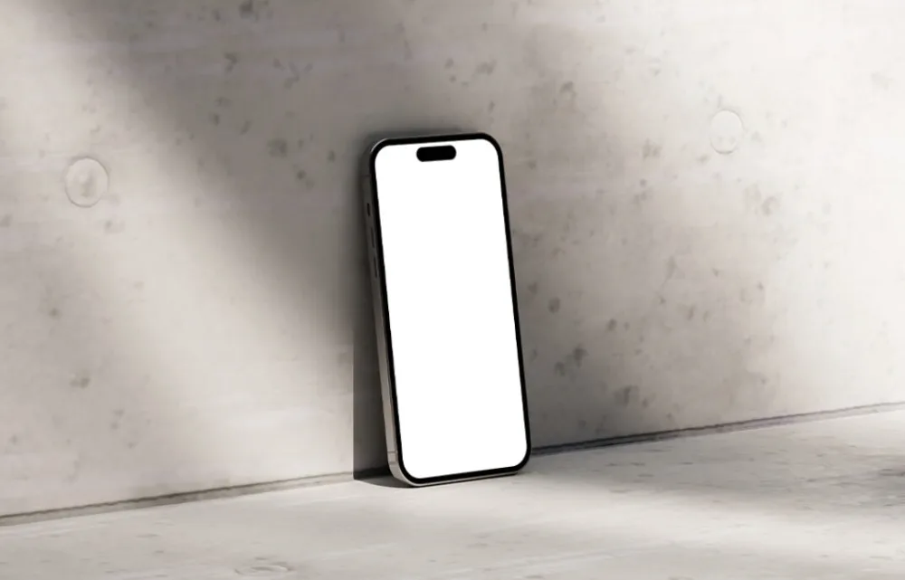 iPhone mockup with a blank white screen, positioned upright against a textured concrete wall. The setting is softly illuminated by natural light, casting subtle shadows on the surface.