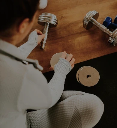 A person wearing white athletic gloves is assembling a pair of dumbbells on a wooden surface, emphasizing a focus on wellness and premium fitness equipment.