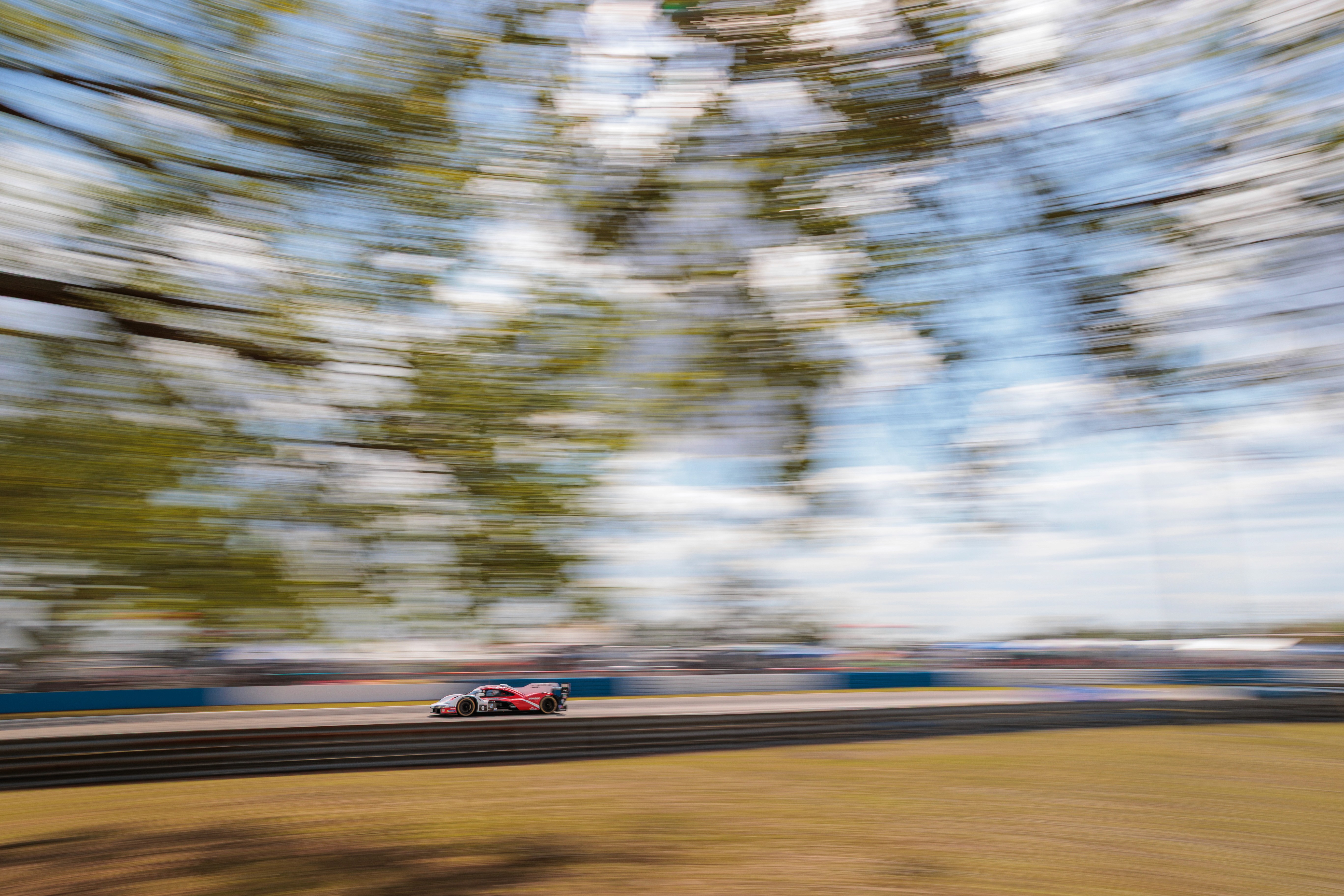 A red Porsche racing car speeds down a track, against a blue sky and trees, with a motion blurred background.