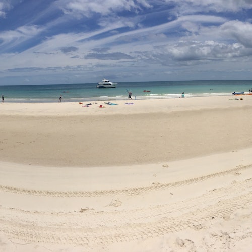 A sandy beach with a few people near the shoreline, a boat on the water, and a partly cloudy sky.