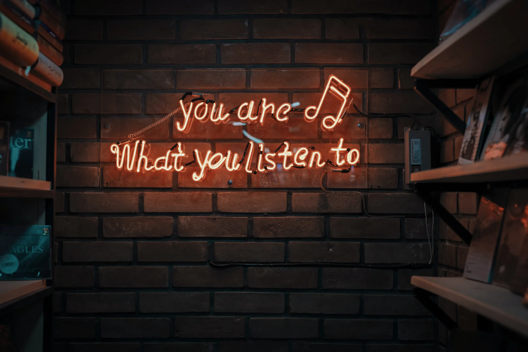 Letreiro de neon com a frase "você é o que você escuta" em uma parede de tijolos