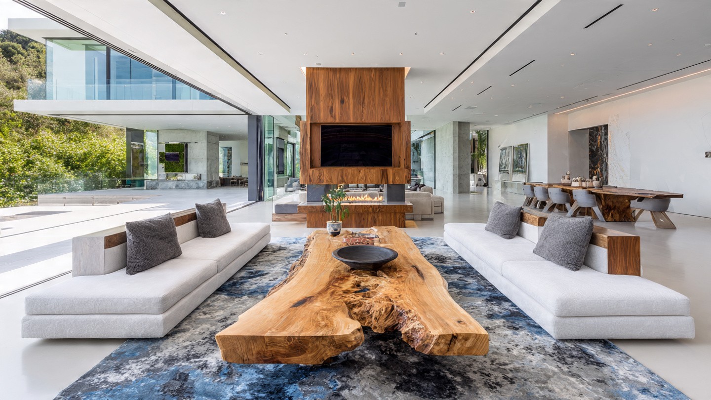 Spacious modern living room with large windows, white sofas, and a polished wood table on a blue rug. A central fireplace adds warmth and elegance.