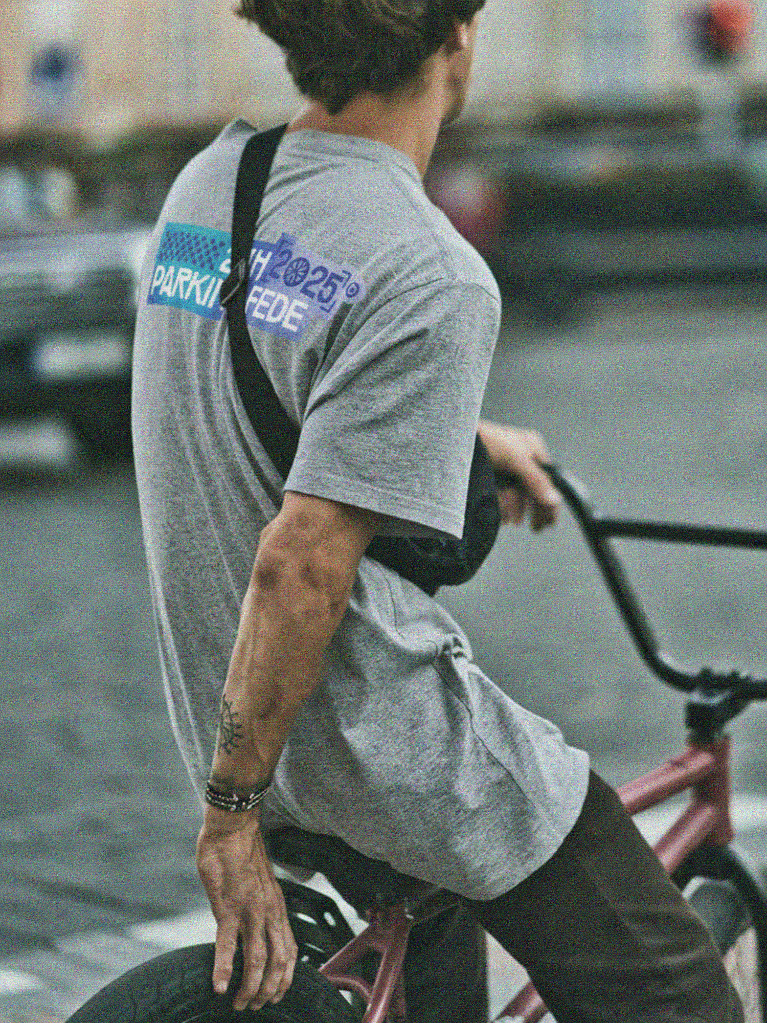 Une prise de vue en conditions urbaines montrant un cycliste de dos, portant un t-shirt gris avec le visuel des 24H Parking Fédé 2025 imprimé en grand format dans le haut du dos. L’image renforce le lien entre l’événement, la culture vélo et un usage terrain, authentique.