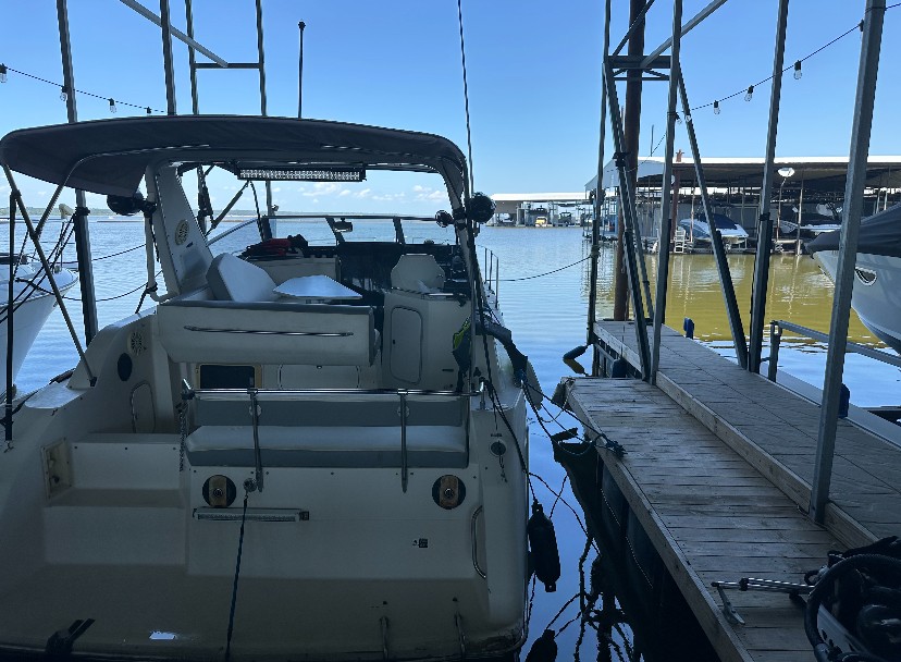 A white motorboat is moored at a wooden dock under a metal canopy, with several other boats and a marina visible in the background.