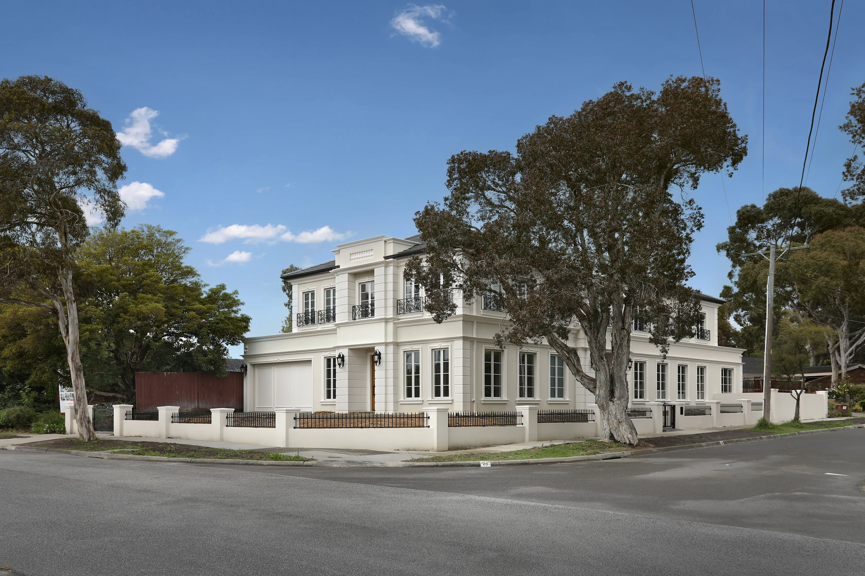 Corner street view of the Wheelers Hill home, a white rendered two-storey residence with classical detailing, mature gum trees, a rendered and wrought-iron fence and a double garage