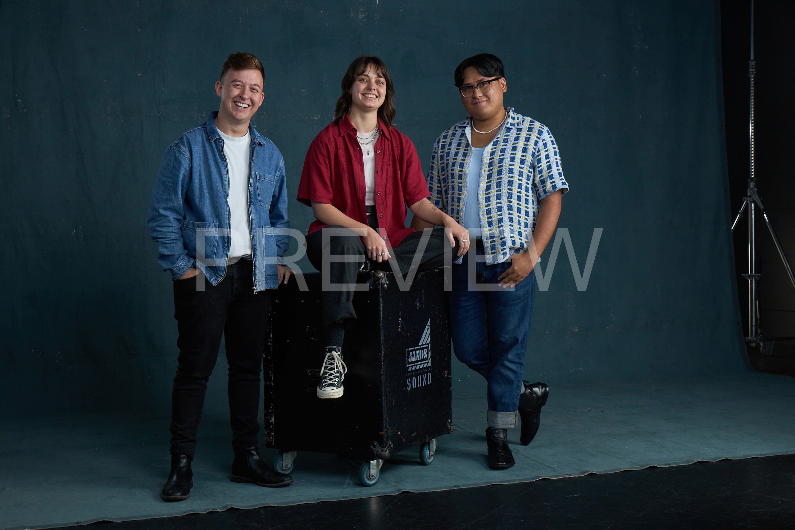 A group portrait of three young adult theatre artists standing and sitting against a dark teal backdrop. A person wearing a navy top and red skirt is seated on a stool in the centre, while two others stand on either side, dressed casually in jeans and T-shirts. All three face the camera with a relaxed, confident expression.