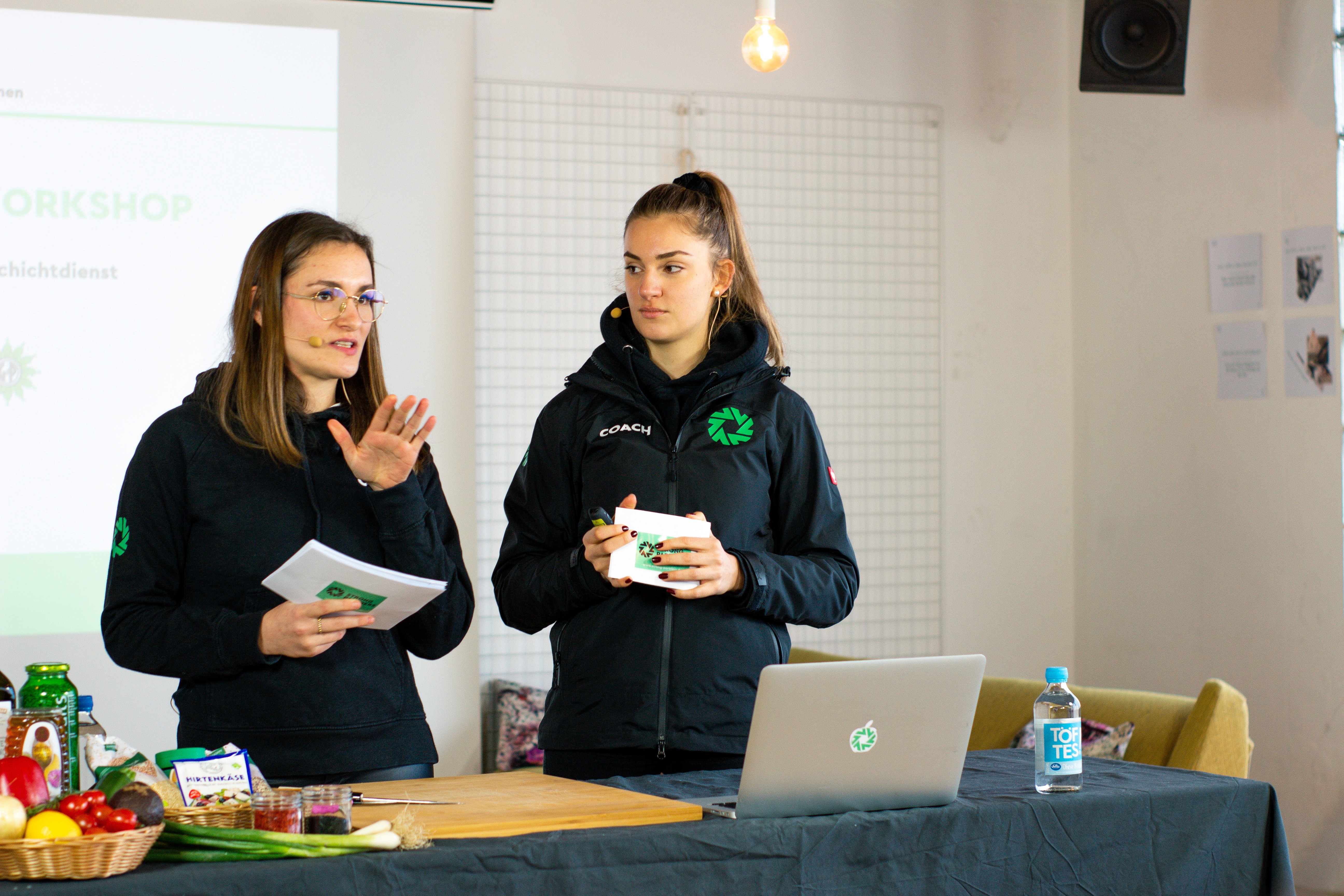 Zwei junge Frauen mit Moderationskarten in der Hand stehen vor einem Tisch mit Lebensmitteln und einem aufgeklappten Laptop, sie sprechen in eine Kamera