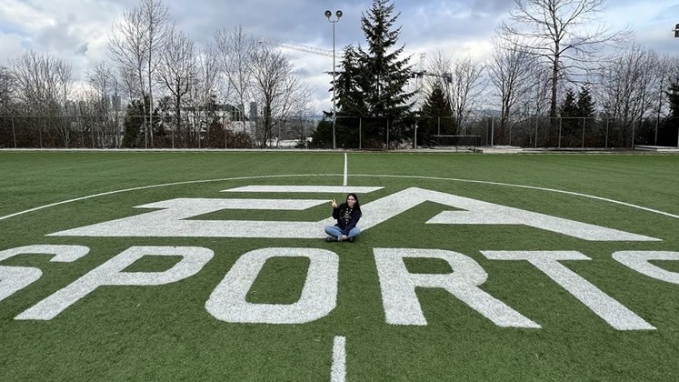Close-up of two professional women in conversation in a contemporary office space. The woman facing the camera has long wavy brown hair, wears a burgundy knit top and nose ring, and displays an engaged, friendly expression. Another woman with short dark hair and pearl earrings is shown from behind. The bright, modern workspace features other colleagues working at desks in the background.
