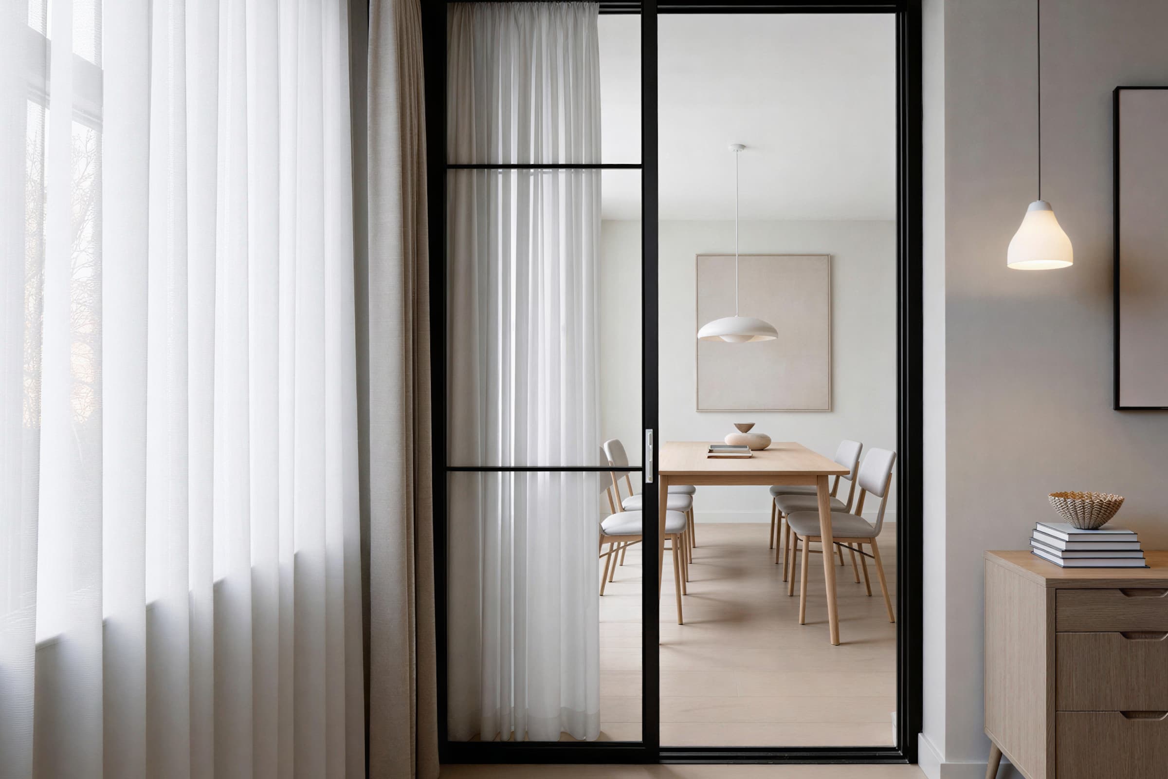 A modern, minimalist dining room features a wooden table with six chairs, illuminated by a sleek pendant light, and separated by a large glass sliding door with black metal frames, enhancing the contemporary design of the bright, softly-lit space.