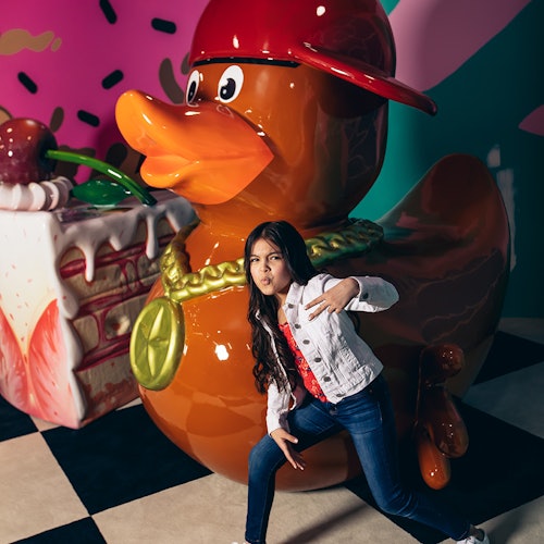 A girl posing playfully in front of a large duck statue wearing a cap, with a giant cake prop in the background.