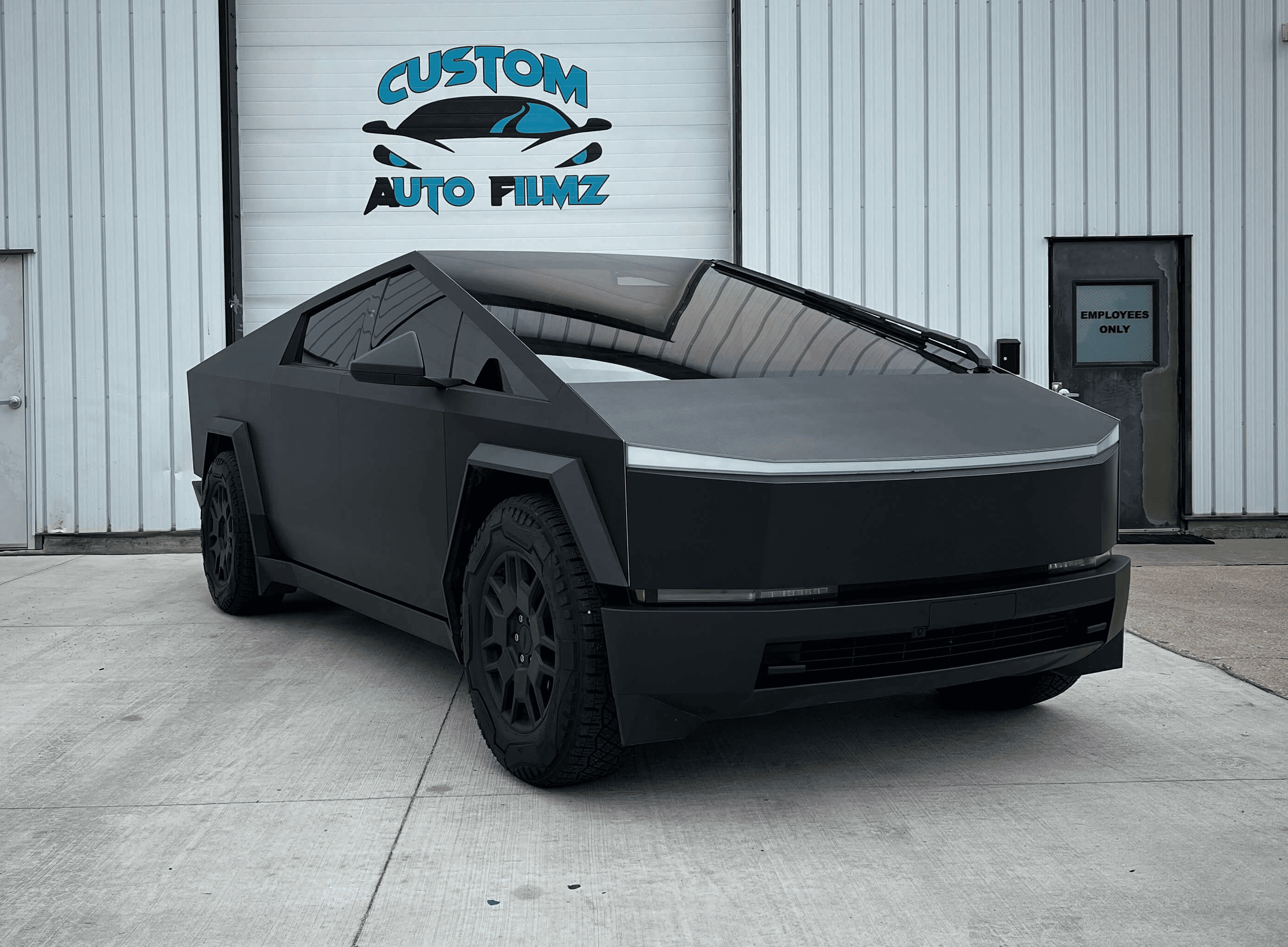 A matte black Tesla Cybertruck is parked on a concrete driveway in front of a white industrial building.