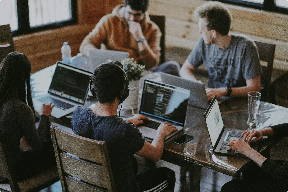 A group of people collaborating around a conference table with laptops in a well-lit, modern workspace.