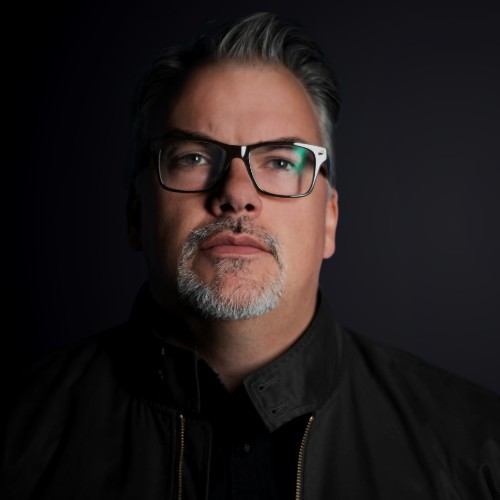 A man with glasses and a brown shirt, crossing his arms.