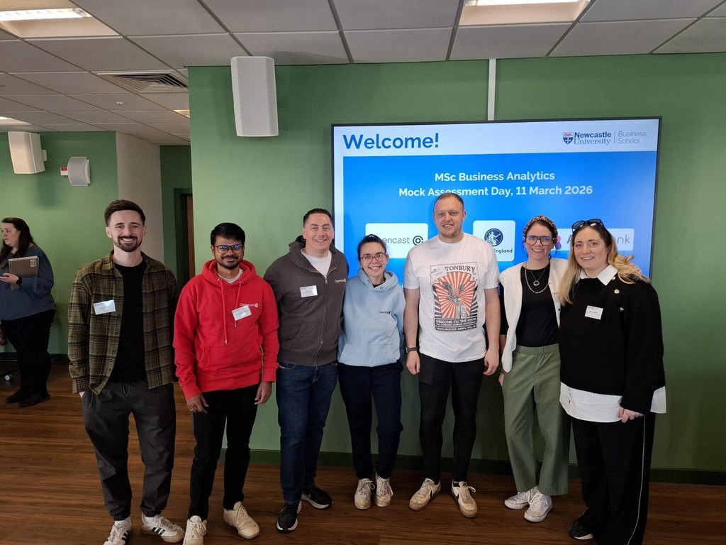 A group of seven people stand in a row in front of a large screen displaying the text “Welcome! MSc Business Analytics Mock Assessment Day, 11 March 2026.” The group is in a room with green walls and wooden flooring. Some individuals wear name badges, and a few laptops and bags are visible in the background, along with other people in the room.