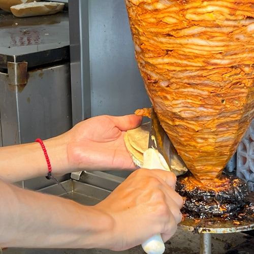 Isang taquero ang naghiwa ng marinated na baboy mula sa vertical na spit papunta sa tortilla, inihahanda ang taco al pastor sa isang tradisyunal na taquería.