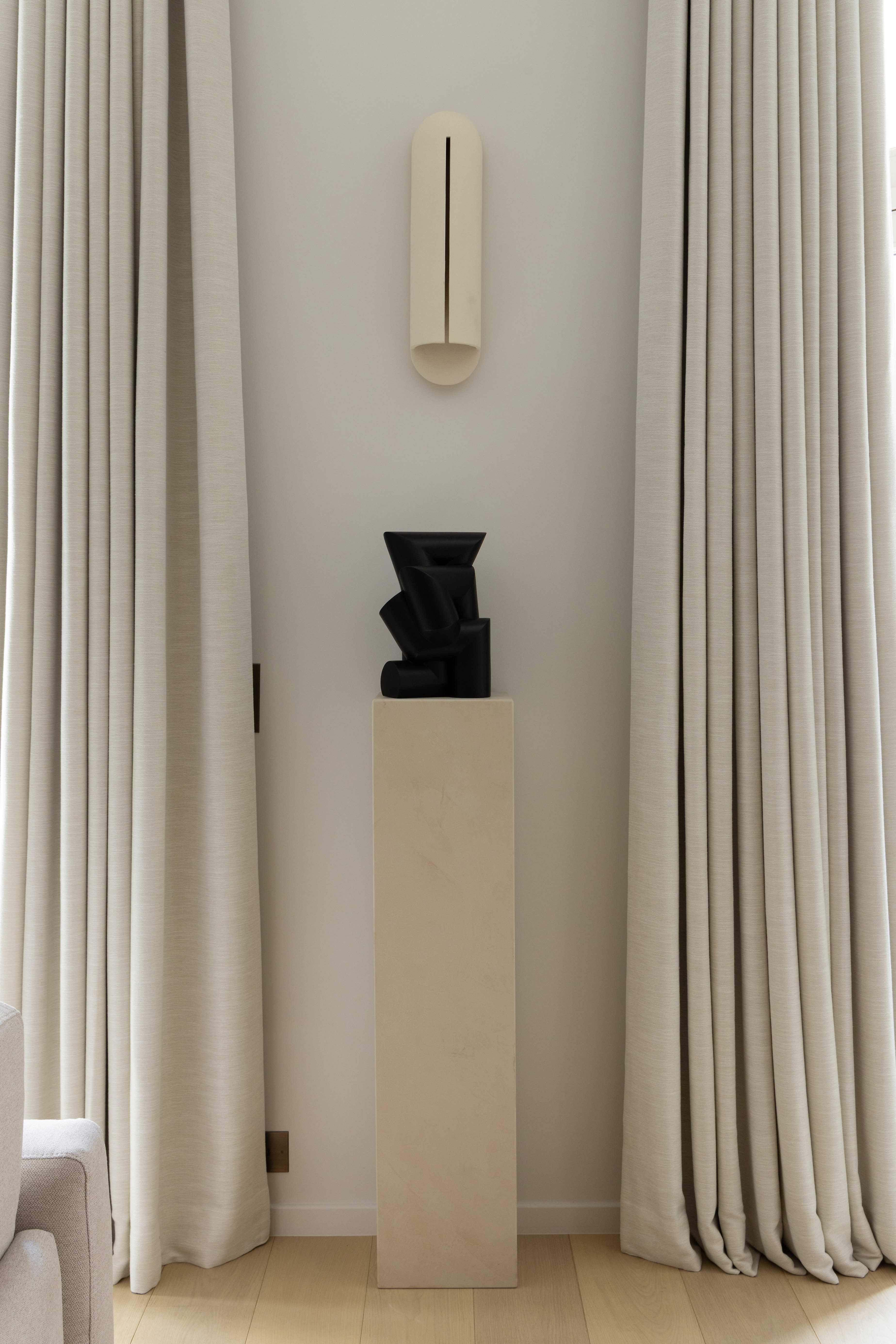 Sculpture noire abstraite sur piedestal beige entre deux rideaux drapes, avec applique murale minimaliste