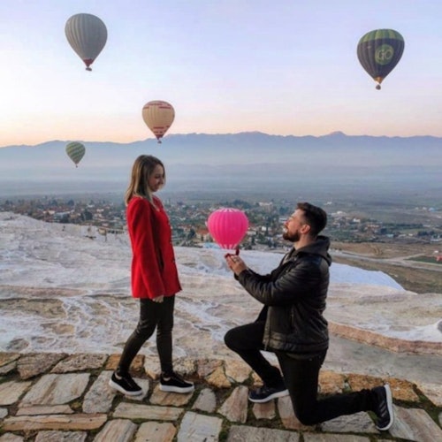Isang lalaki ang nakaluhod habang hawak ang rosas na lobo, nagpo-propose sa isang babaeng naka-pulang coat, habang may mga lumilipad na hot air balloons sa kalangitan.