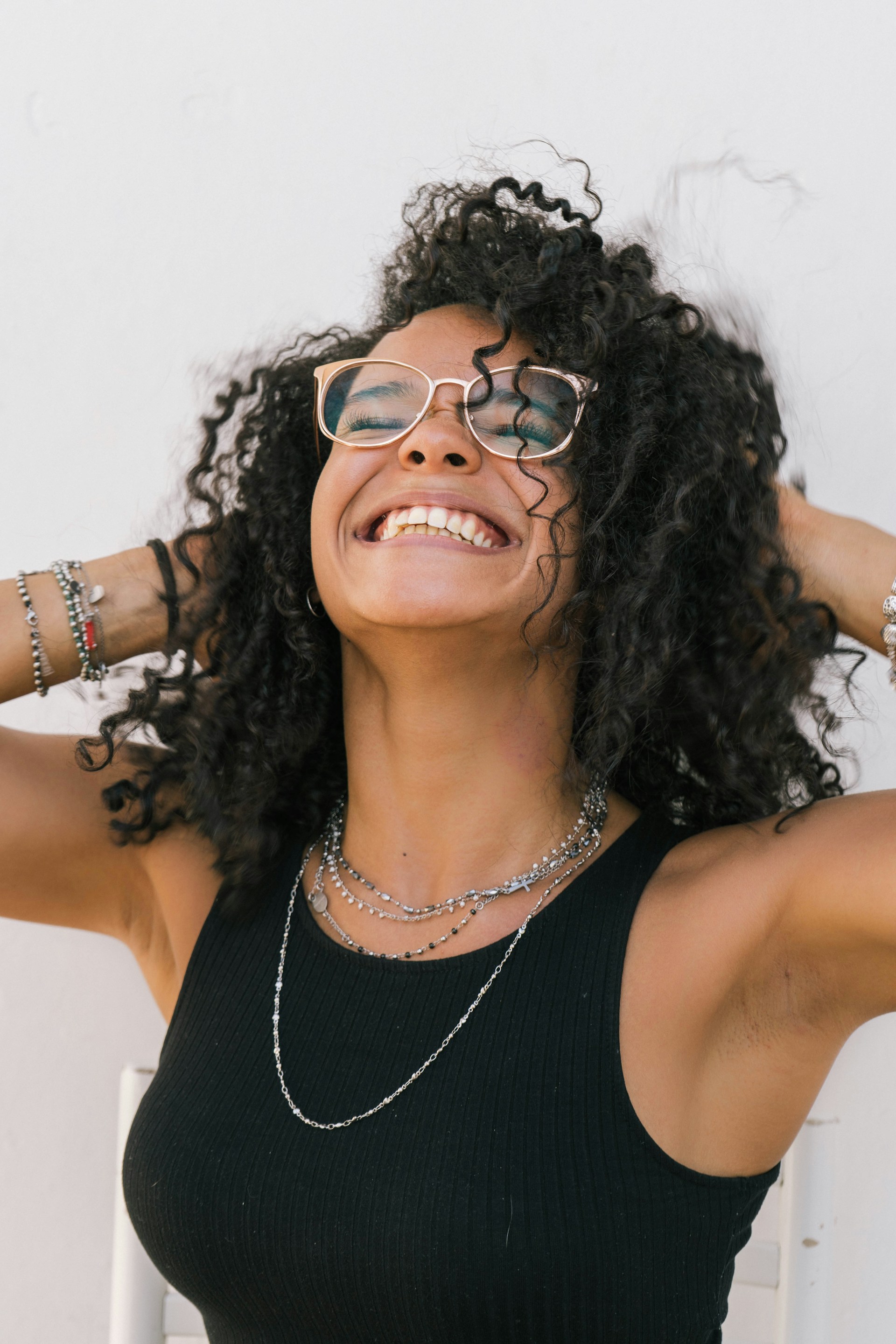A woman with voluminous dark curls and glasses laughing with her head tilted back, wearing a black tank top and silver necklaces.