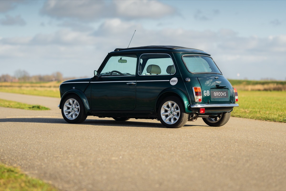 A classic green Mini Cooper parked on a road, surrounded by an open landscape and blue sky.