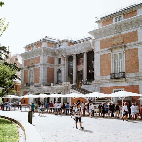 מבנה לבנים אדומות עם עמודים ופוסטר גדול, כאשר אנשים עומדים בתור תחת מטריות מלפנים, וקצת ירק משמאל.