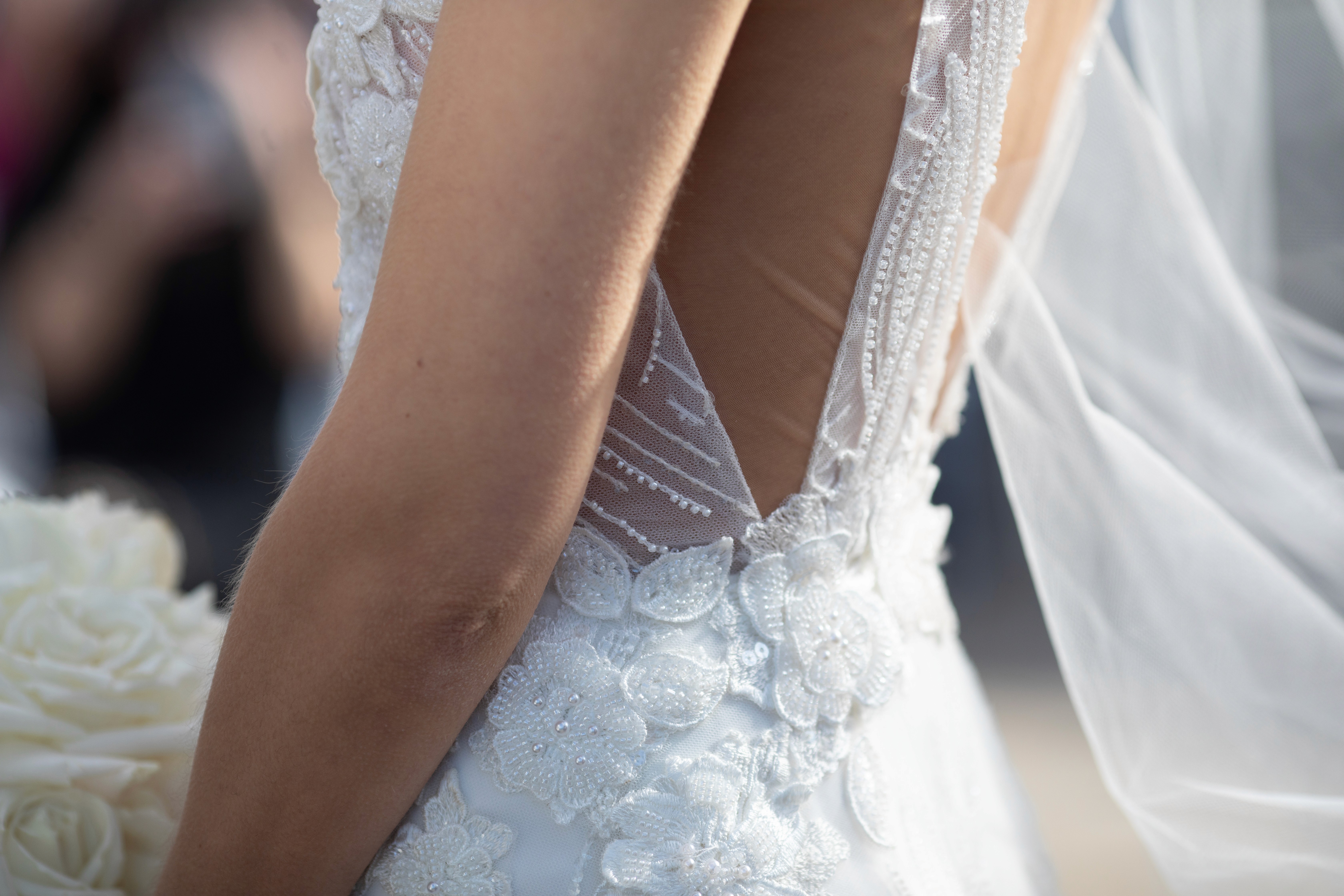 Wedding photographer – bride in elegant gown standing by window with flowing curtains and bouquet
