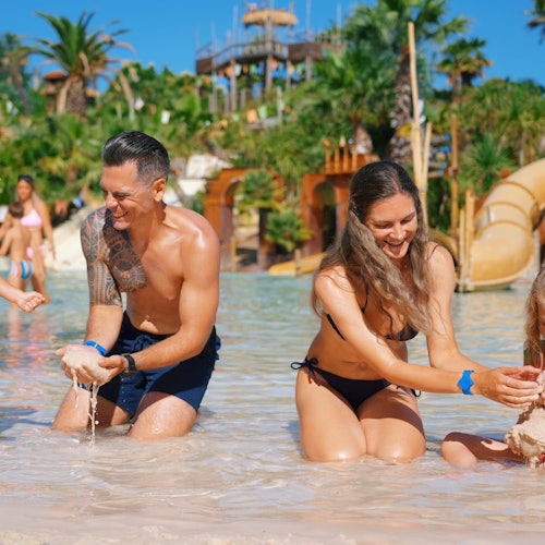 A joyful family playing in shallow water at a water park, with slides and lush greenery in the background.
