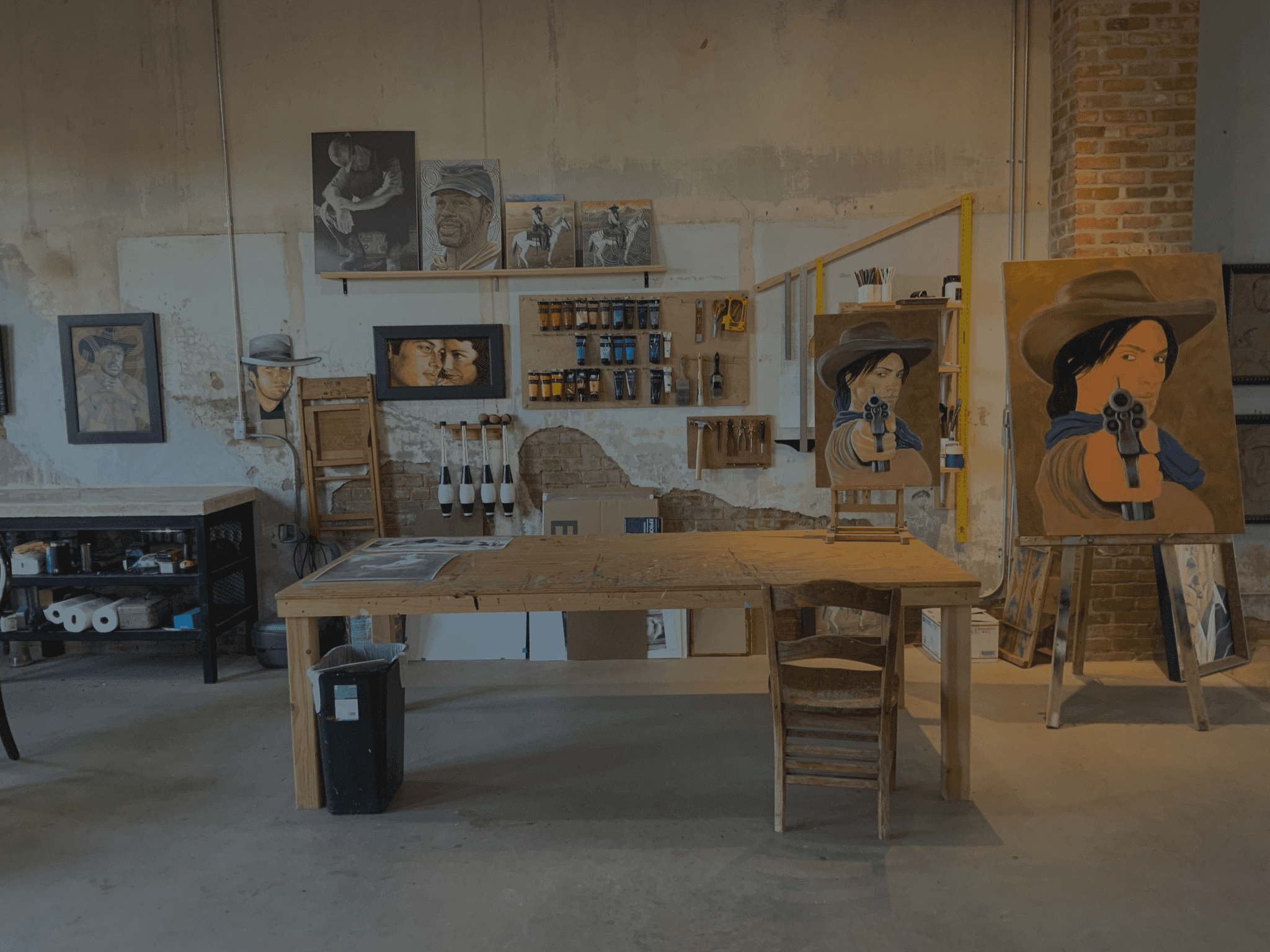 Chuck's workspace with a large table covered in brushes, paint, and masonite with several paintings on the wall and one on an easel next to the table.