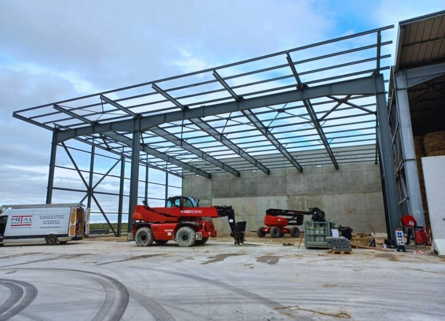 Structure métallique en construction sur sol en gravier, avec nacelles élévatrices en action sous un ciel bleu.