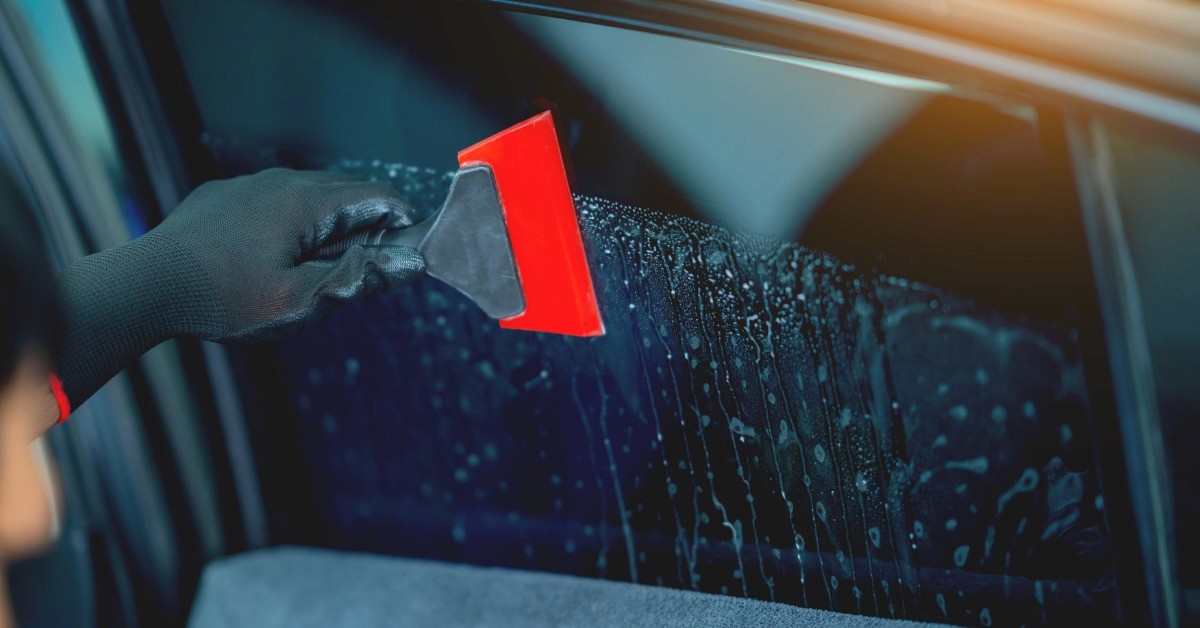 A black gloved hand using a red squeegee on a wet tinted car window with dark trim and a gray towel below.