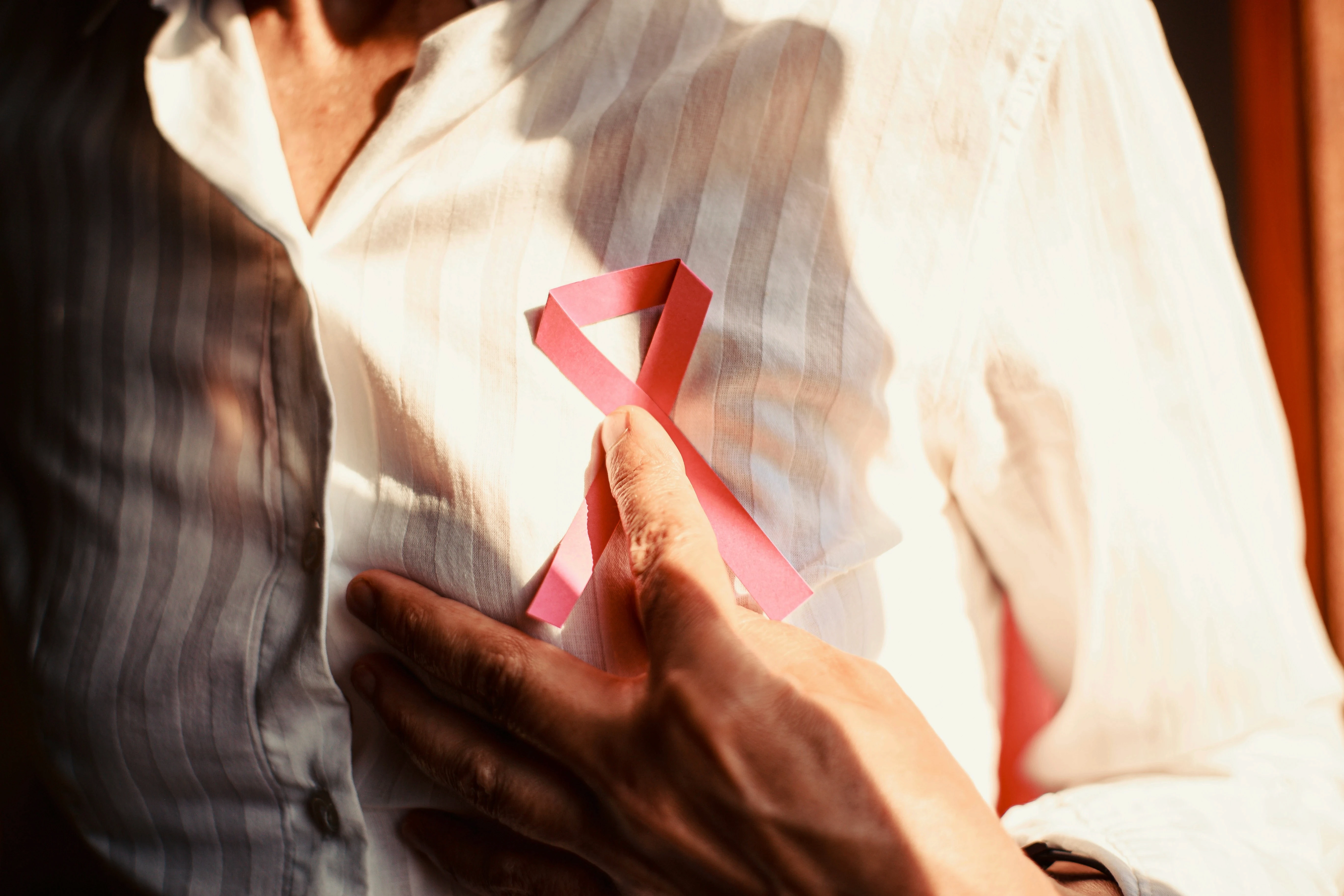 Person holding a red HIV/AIDS awareness ribbon against their chest, symbolizing survival, collective trauma, and the lasting mental health impact of the AIDS crisis on the queer community.