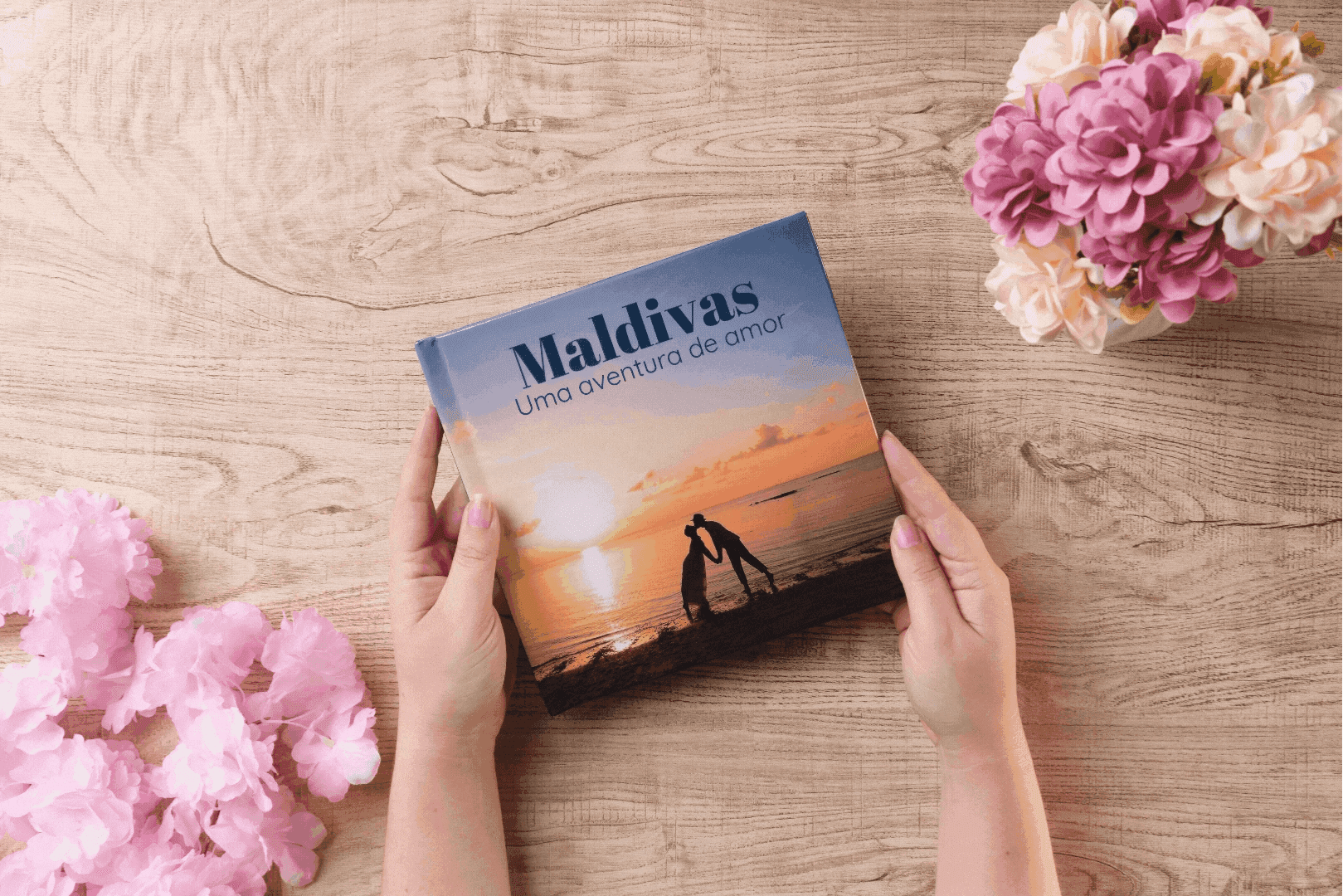 Mãos segurando fotolivro com capa personalizada das Maldivas, com casal ao pôr do sol e flores decorativas ao redor, sobre mesa de madeira clara
