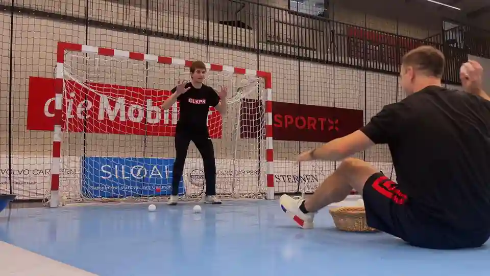 Handball goalkeeper facing a seated coach holding a small white ball during a reaction drill in front of the goal.