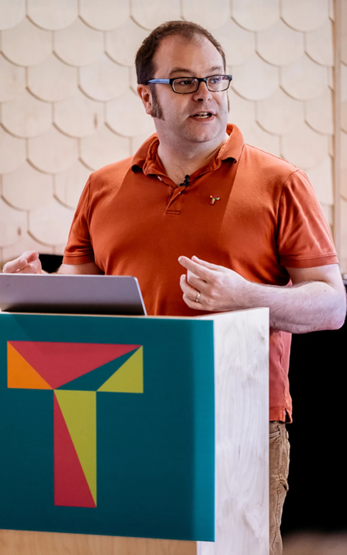 Person wearing an orange polo shirt stands behind a podium with a colorful geometric logo, holding a laptop and gesturing with hands.