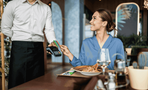 A man in a white shirt hands a receipt to a woman in a blue shirt at a restaurant table with food and drinks.