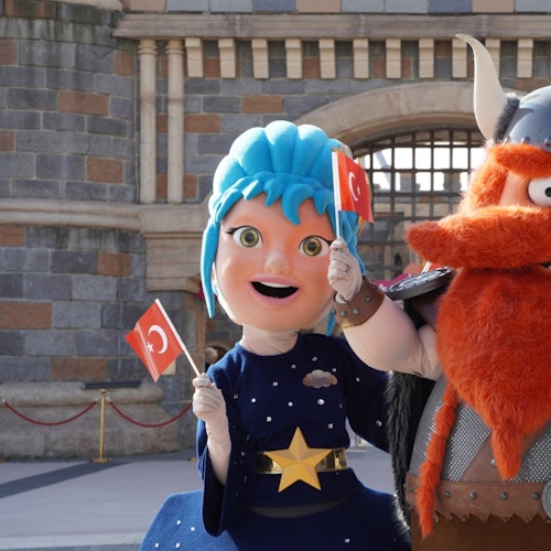 Dalawang tauhan na may kasuotan, ang isa ay may asul na buhok at ang isa naman ay nakabihis ng berserk, ay may hawak na mga bandila ng Turkey sa harap ng isang gusaling bato.