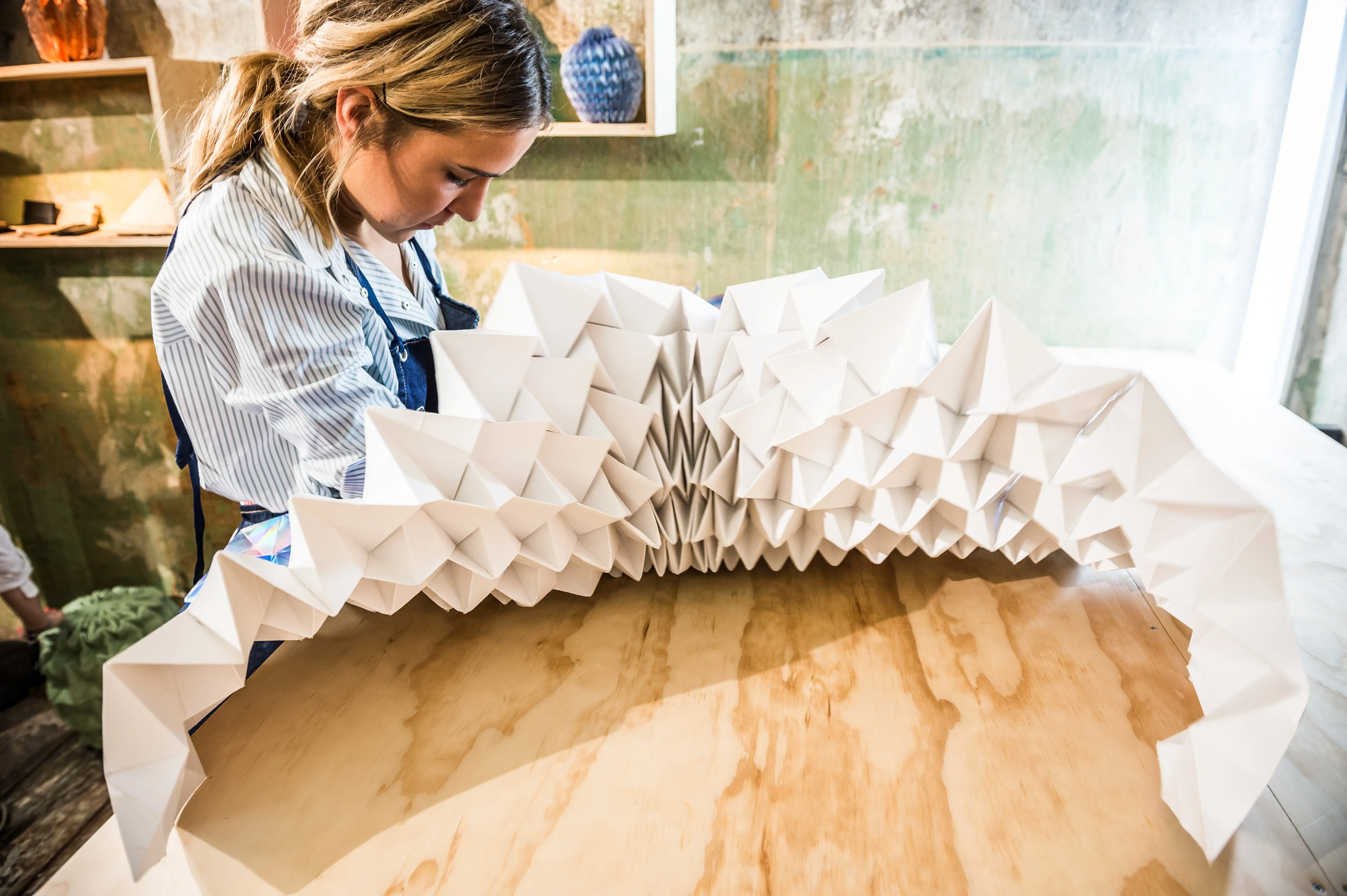 Woman folding a large white origami paper sculpture on a plywood table at the MUSTANG Street ART Lab.