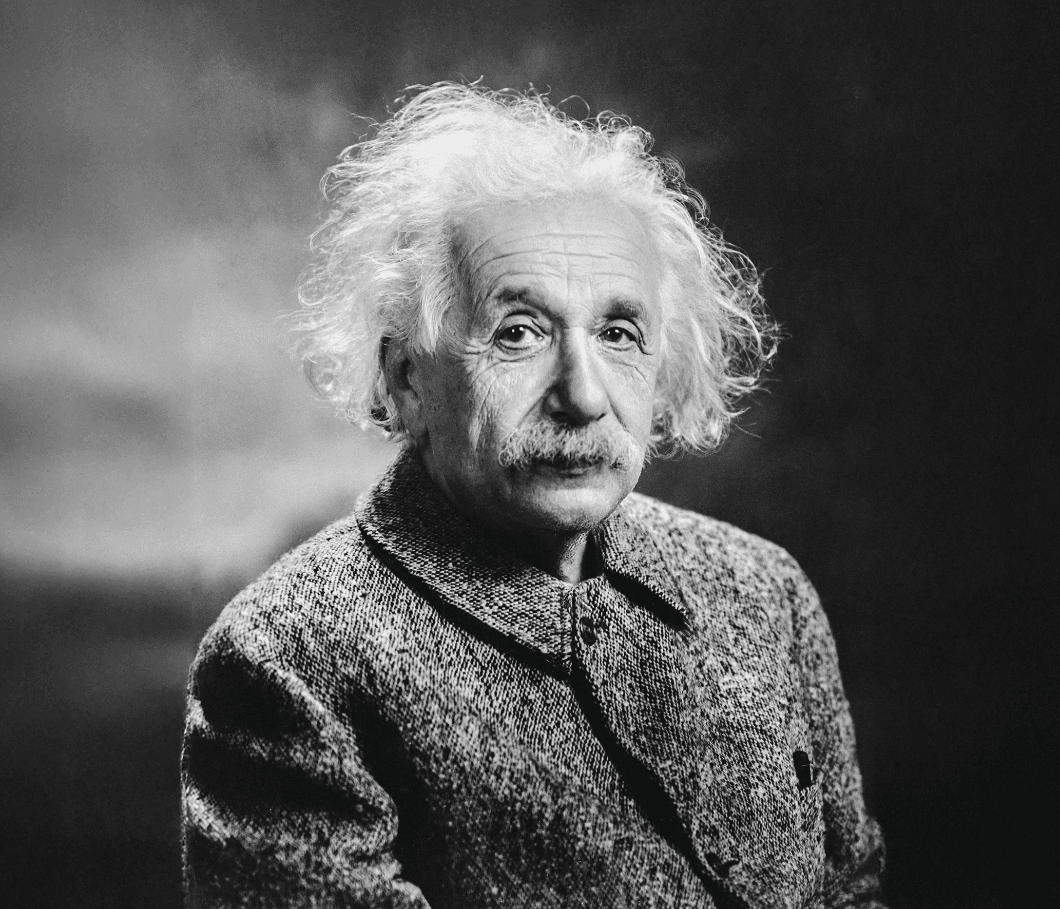 Black-and-white portrait of an elderly man with wild white hair and a thick mustache, wearing a textured jacket and gazing calmly toward the camera against a softly blurred background.