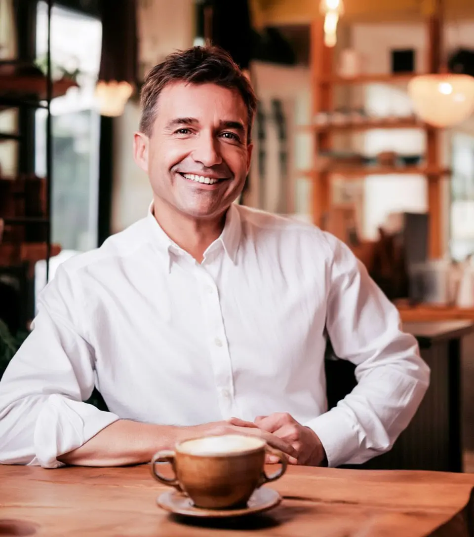 Man in white shirt smiling with a latte on wooden table.