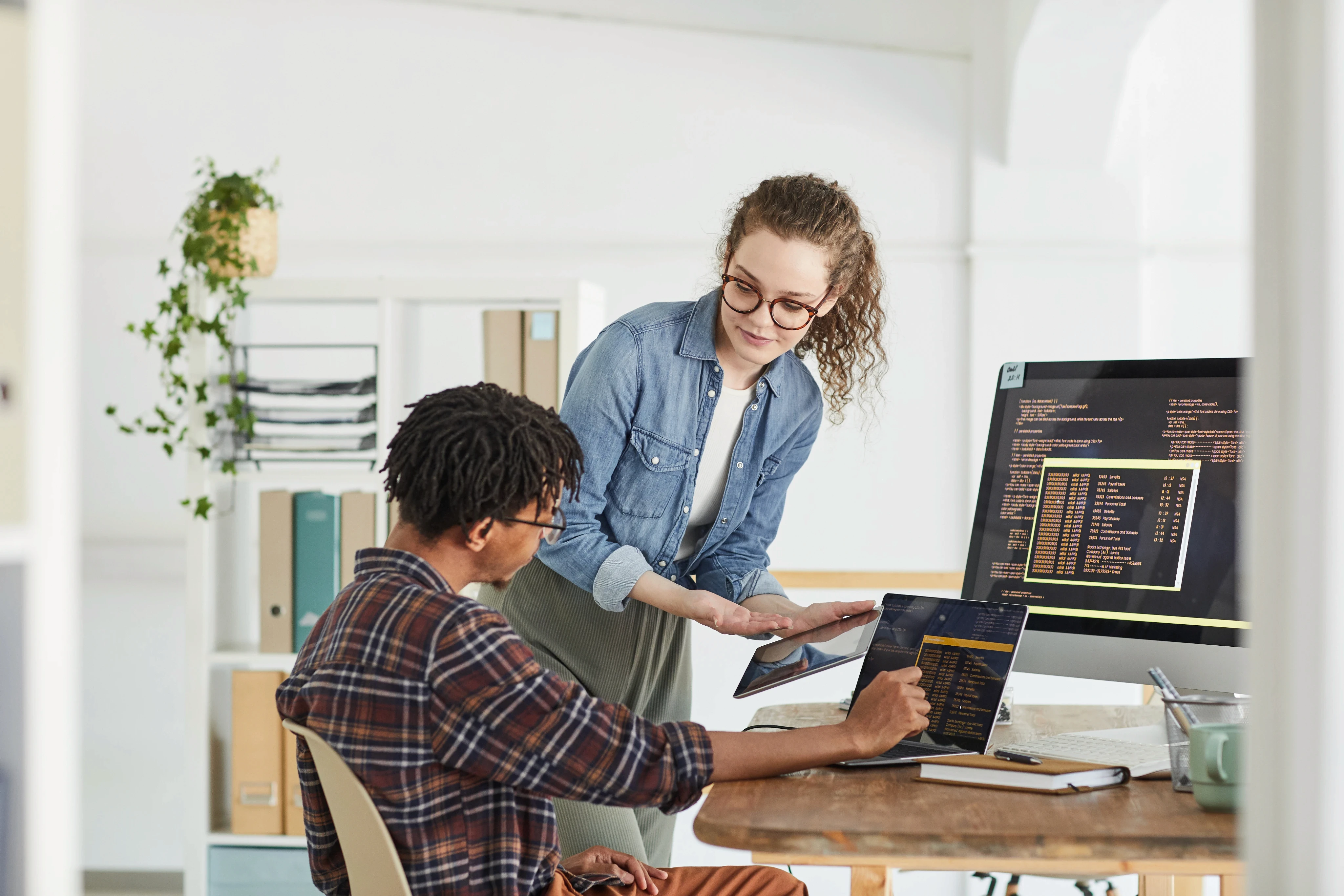 Two software developers collaborating on code shown on a desktop monitor and laptop in a bright, modern office setting.