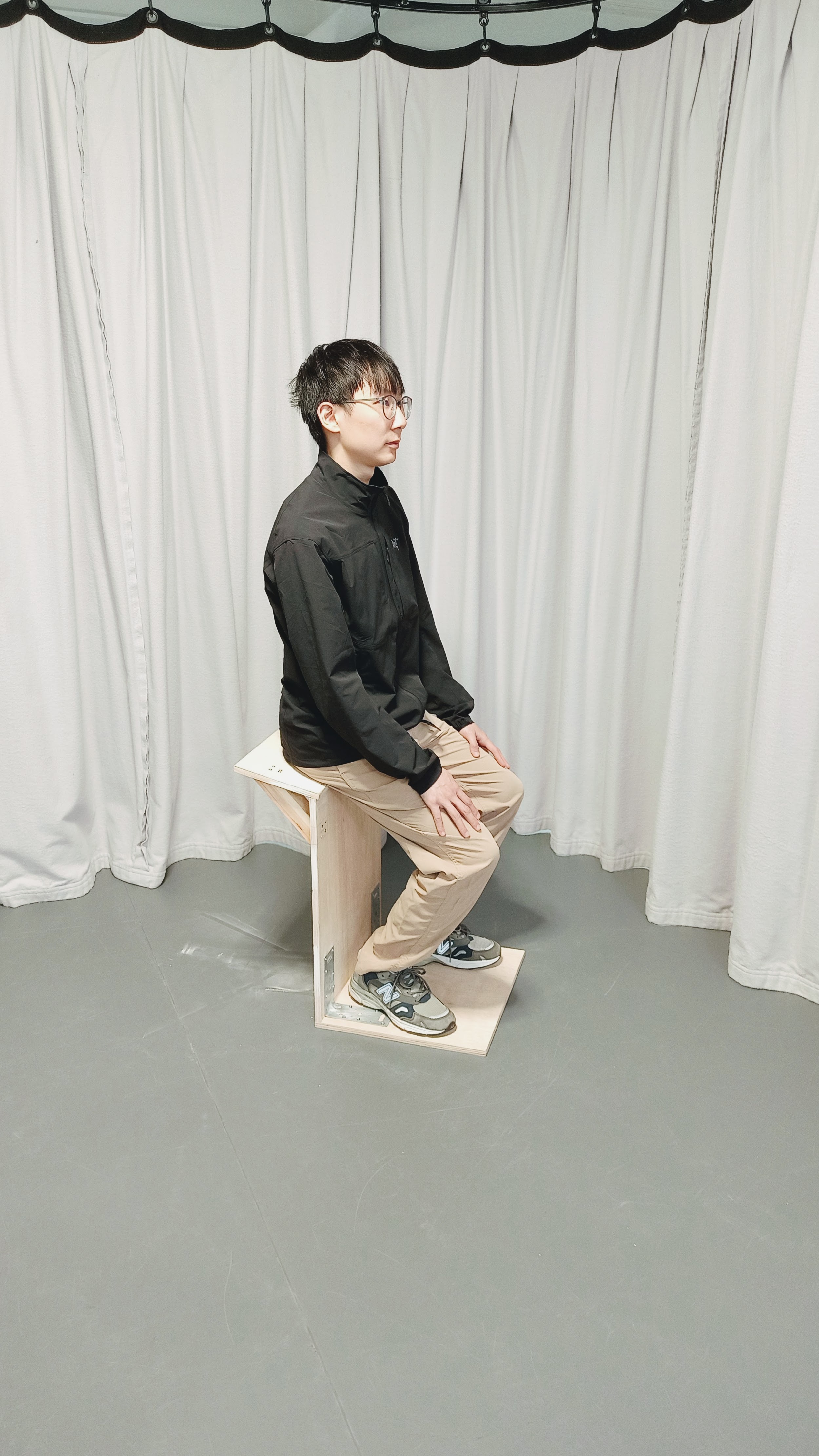 Person sitting upright on a compact plywood chair in front of white curtains.