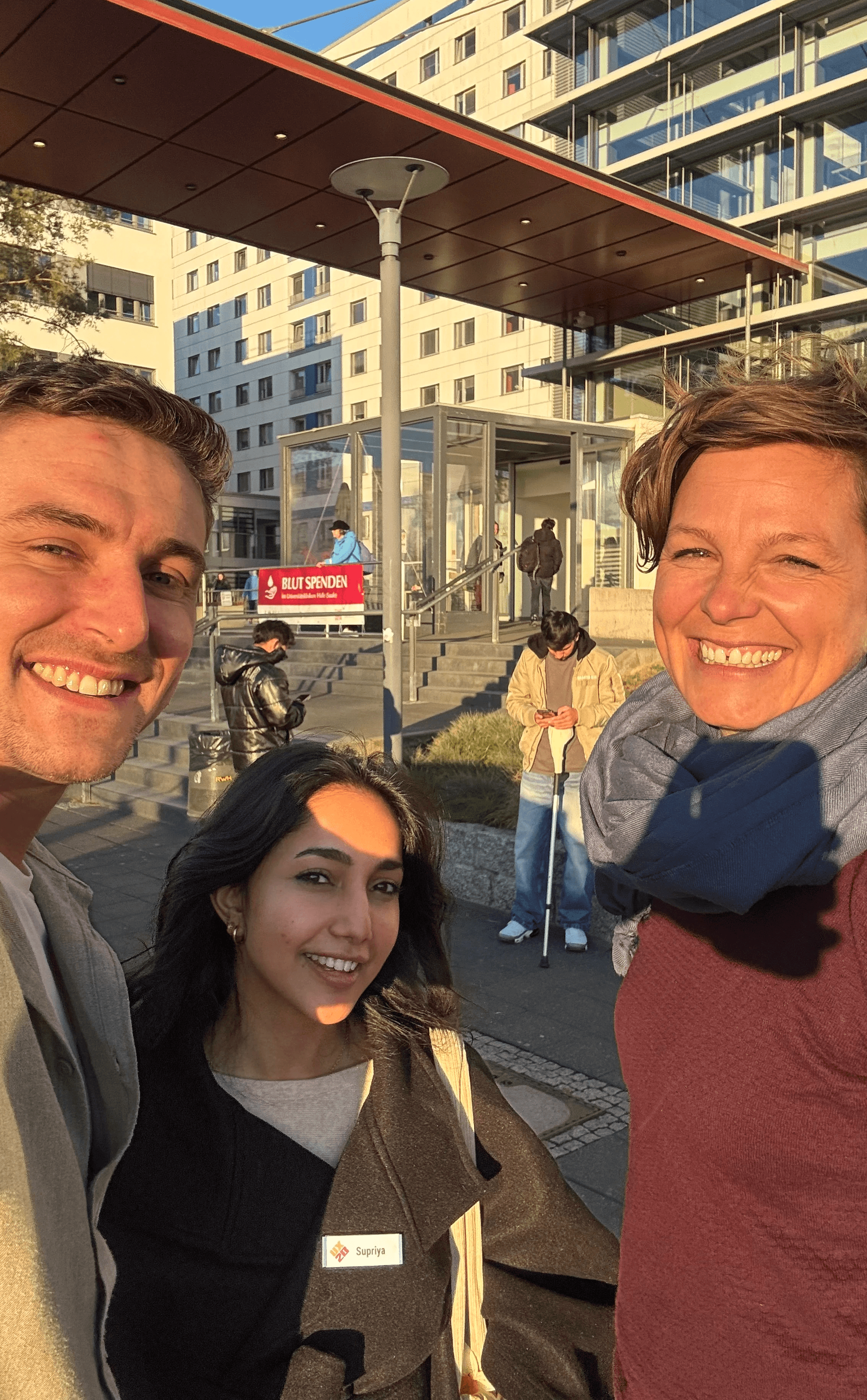 Drei Personen mit Namensschildern stehen vor einem modernen Gebäude mit Glasfassade und überdachtem Eingangsbereich, im Hintergrund weitere Personen und ein Schild mit rotem Hintergrund.