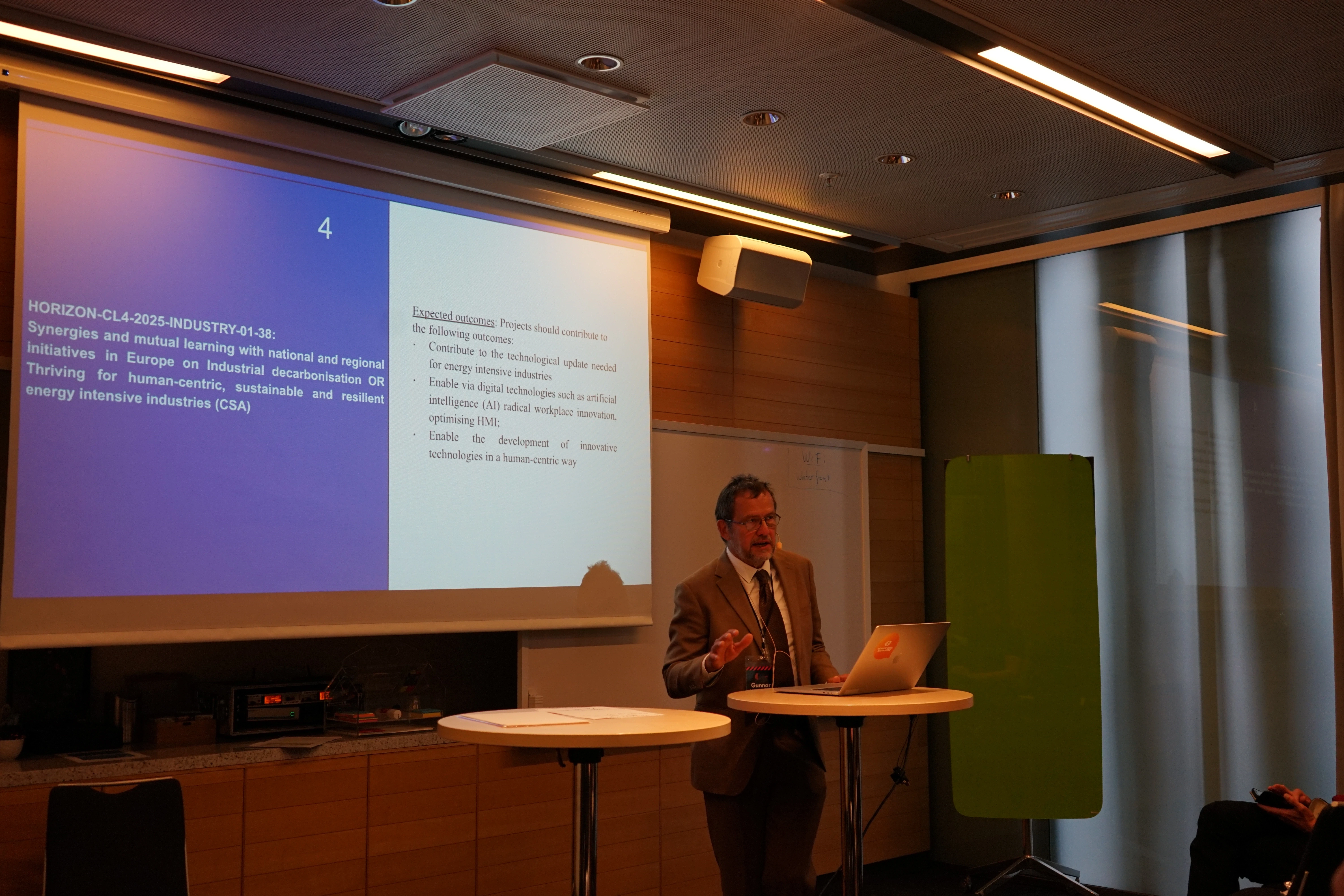 Presenter speaking during a Horizon Europe workshop, standing beside a projected slide on expected outcomes for industrial innovation