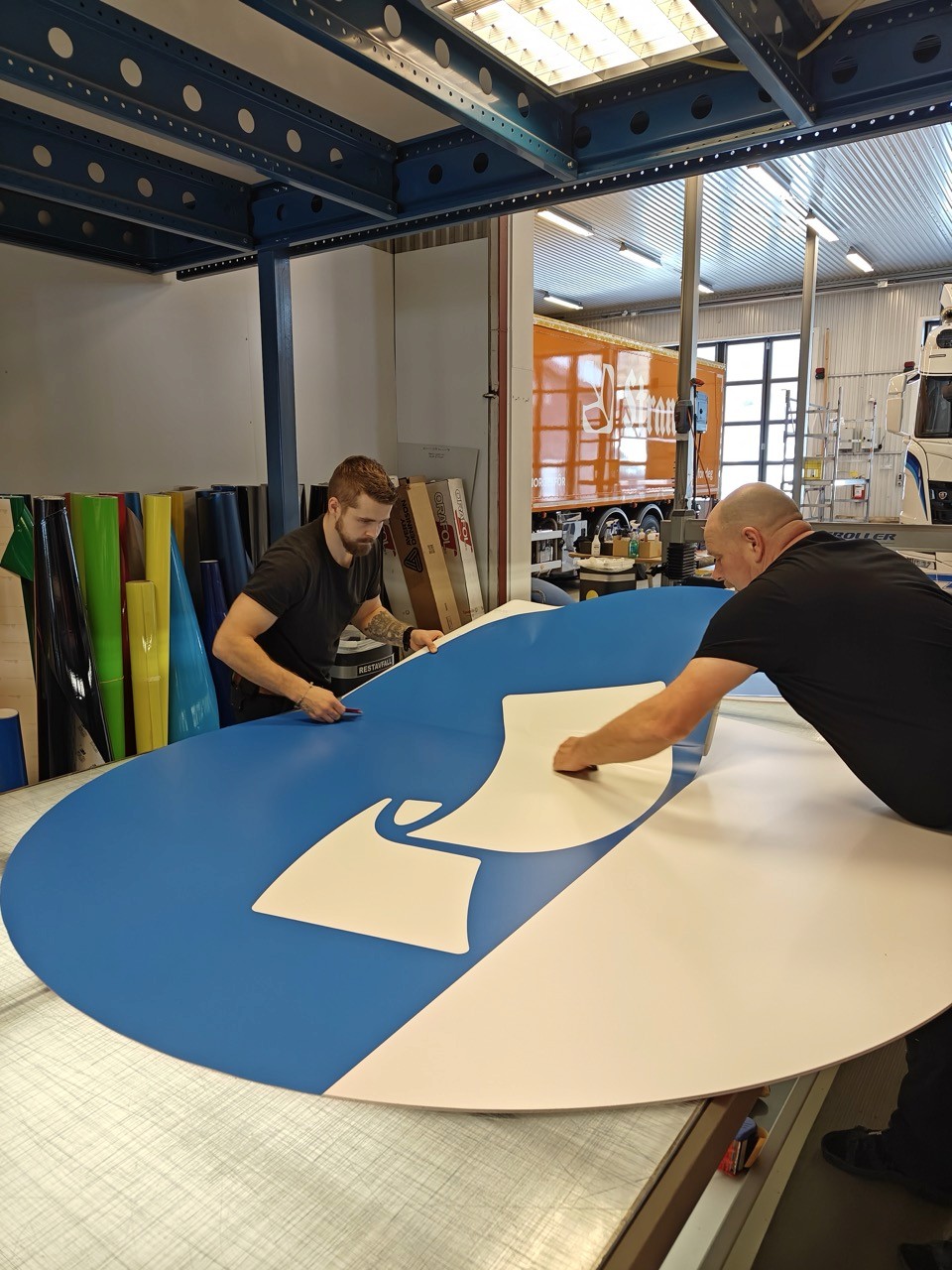 To personer jobber sammen med å kutte et stort blått og hvitt ark med materiale i et moderne industrimiljø, med fargerike ark og verktøy synlige i bakgrunnen, noe som fremhever samarbeid og presisjon i en produksjonssetting.