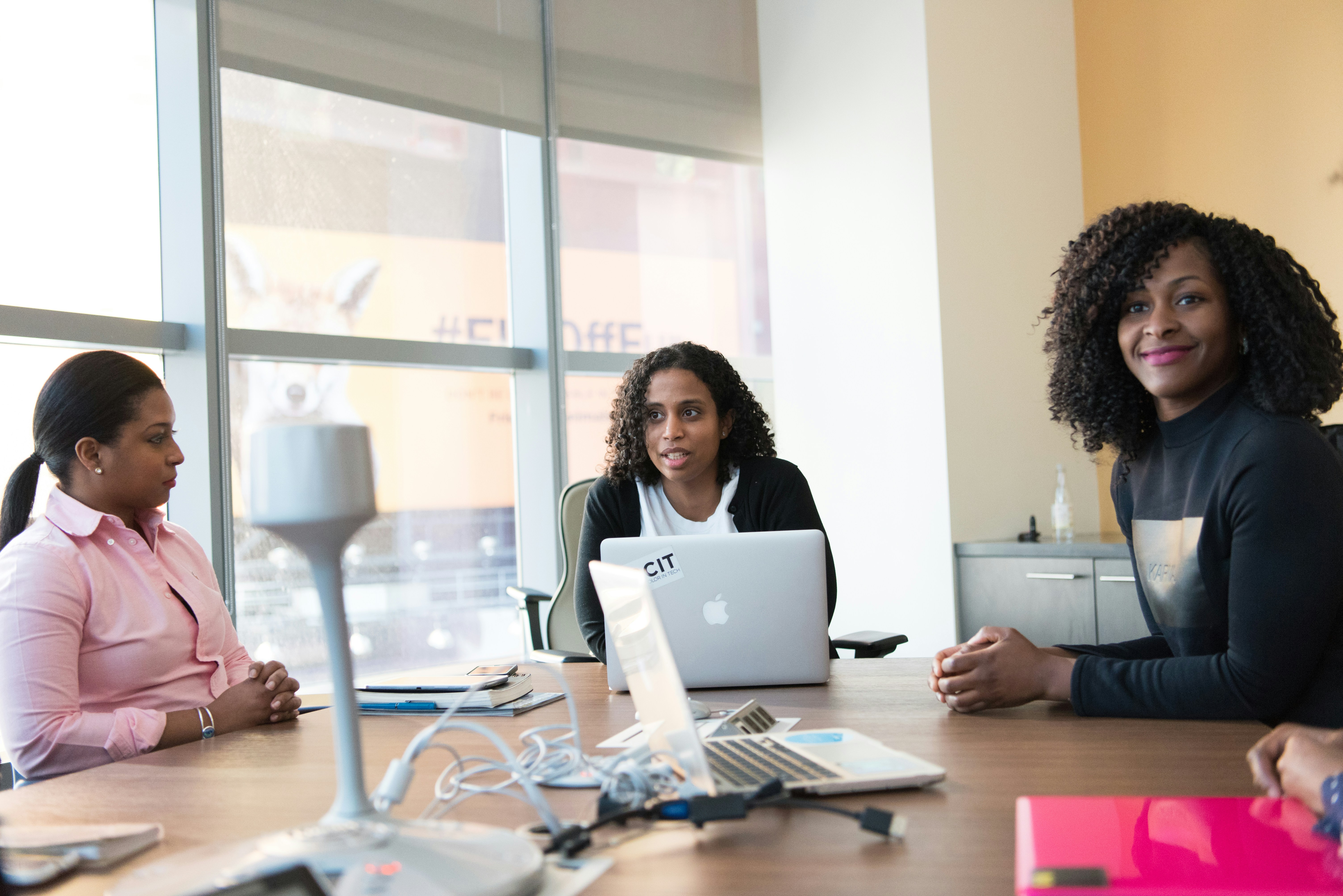 Three professional women in a meeting, one speaking with a MacBook open in front of her, in a modern office environment with natural light.